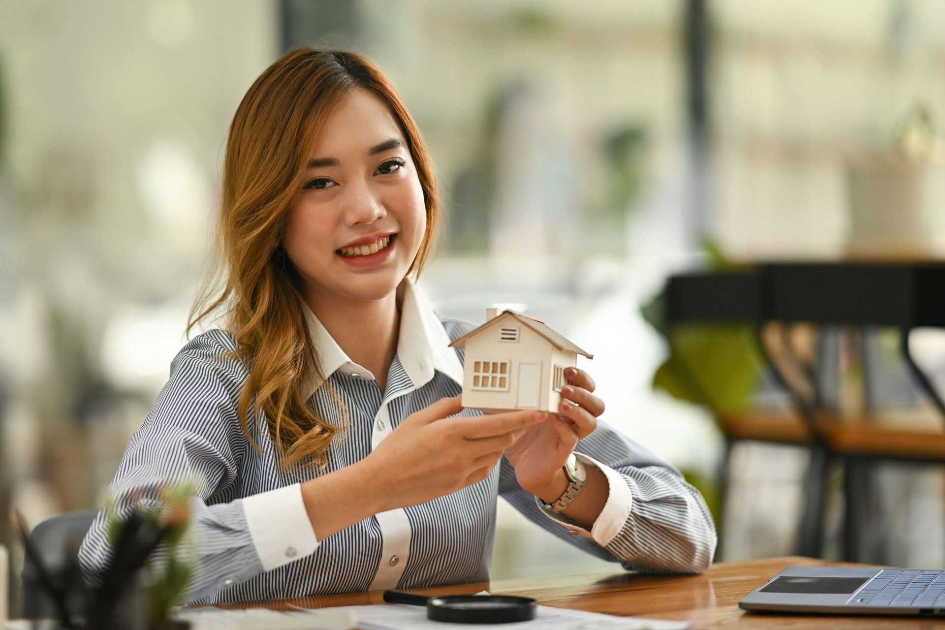 Une femme professionnelle souriante tenant une petite maison modèle symbolise l’immobilier, l’investissement immobilier, l’accession à la propriété, la vente de propriétés, le conseil immobilier ou les opportunités d’investissement